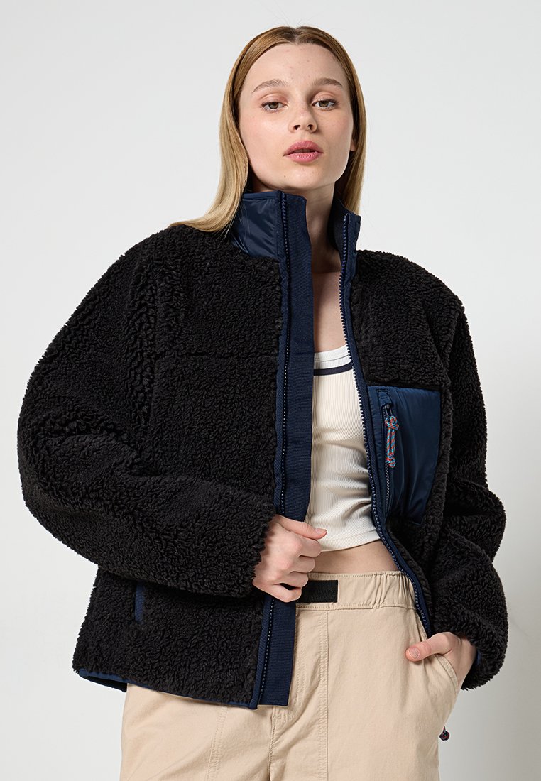 Schwarze Fleecejacke mit hohem Kragen, strukturiertem Stoff, durchgehendem Reißverschluss und einer blauen Nylonbrusttasche mit einem bunten Reißverschlussanhänger.