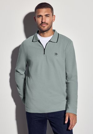 Mann mit kurzen Haaren und Bart, der ein hellgrünes Zip-Kragen-Langarmhemd über einem weißen T-Shirt und dunklen Jeans trägt und vor einem schlichten Hintergrund steht.