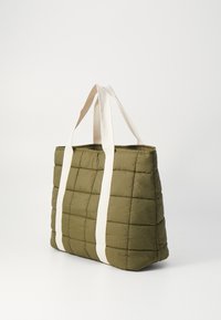 Grønn foret totebag med hvite håndtak, med rektangulær form og polstret tekstur. Merkbar rutenett-mønster på overflaten.