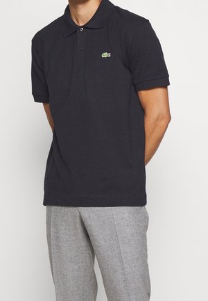 Homme portant un polo noir à manches courtes avec un petit logo crocodile vert, associé à un pantalon gris clair, sur un fond blanc.