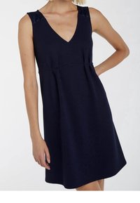 Mouwloze marineblauwe textuur jurk met V-hals en getailleerde taille, gedragen door iemand met één hand op de heup.