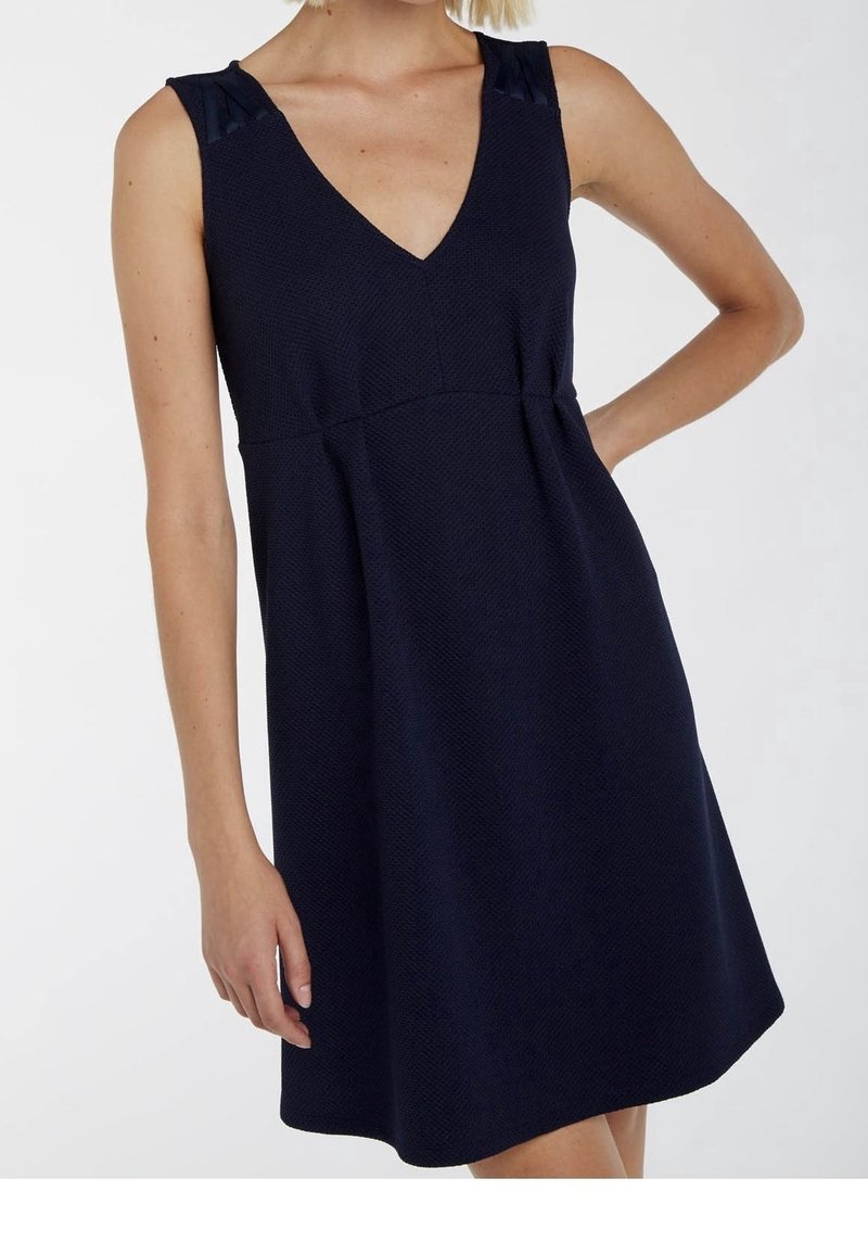 Mouwloze marineblauwe textuur jurk met V-hals en getailleerde taille, gedragen door iemand met één hand op de heup.