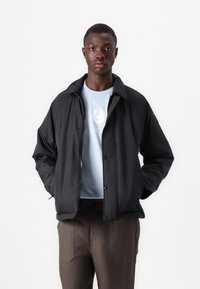 Black padded jacket with a high collar, snap buttons, and two side pockets, worn over a light blue t-shirt and brown pants.