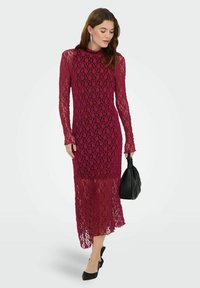 Robe longue en dentelle bordeaux avec manches longues, col montant et motif en maille ajourée. Accessoirisée d'un sac à main noir et de chaussures à bouts pointus.