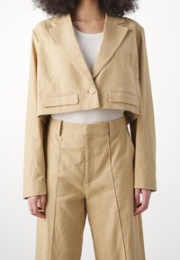 Przycięty beżowy blezer z teksturowanej tkaniny, pojedyncze zapięcie na guzik i dwie przednie kieszenie, w zestawie z beżowymi spodniami z wysokim stanem.