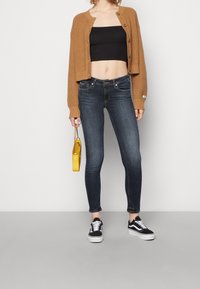 Cárdigan cropped marrón sobre un top corto negro, combinado con jeans ajustados azul oscuro y zapatillas negras. Se sostiene un pequeño bolso amarillo.
