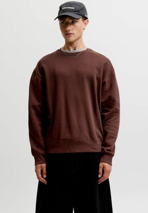 Jeune homme portant un sweat-shirt marron foncé, un pantalon noir, une casquette noire Jack & Jones et un fin collier chaîne, debout devant un fond uni.