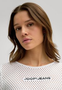 Weißer mesh gestrickter Pullover mit dem schwarz gestickten "JOOP JEANS" Logo. Nahaufnahme des Gesichts des Models mit schulterlangen welligen braunen Haaren.