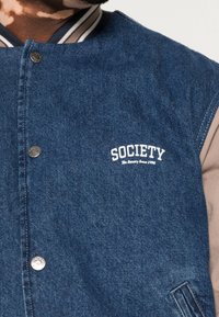 Giacca di denim blu scuro con maniche grigie, caratterizzata da un logo ricamato bianco "Society", bottoni a pressione e profili a righe sul collo.
