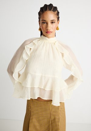 Kvinde med fletninger iført guld øreringe, en gennemsigtig hvid bluse med flæser og et gulbrunt ternet skørt.