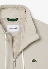 Beige leichte Jacke mit einem Mesh-Futter, ausgestattet mit einem Frontreißverschluss, einer Kordelzugkapuze und einem kleinen grünen Krokodil-Logo auf der Brust.