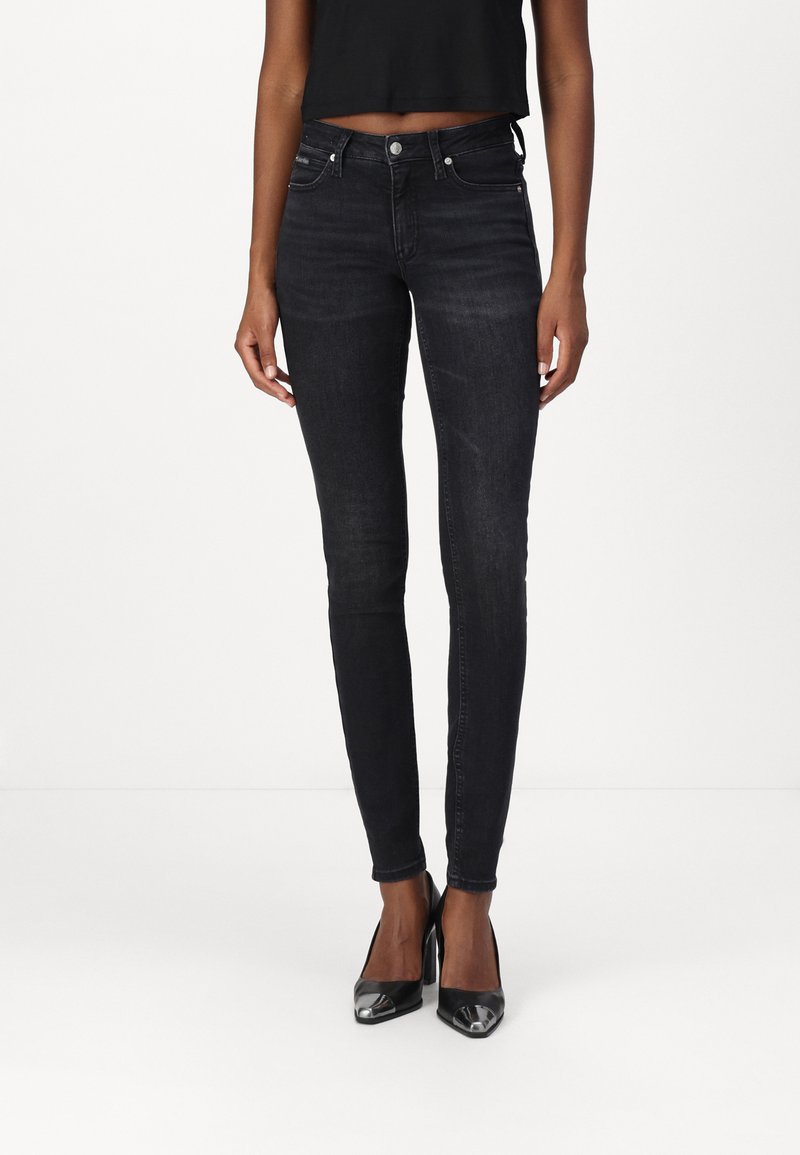 Jeans negros ajustados de mezclilla con una textura suave, diseño de cinco bolsillos y detalles en hardware plateado. Combinados con zapatos de tacón alto negros.