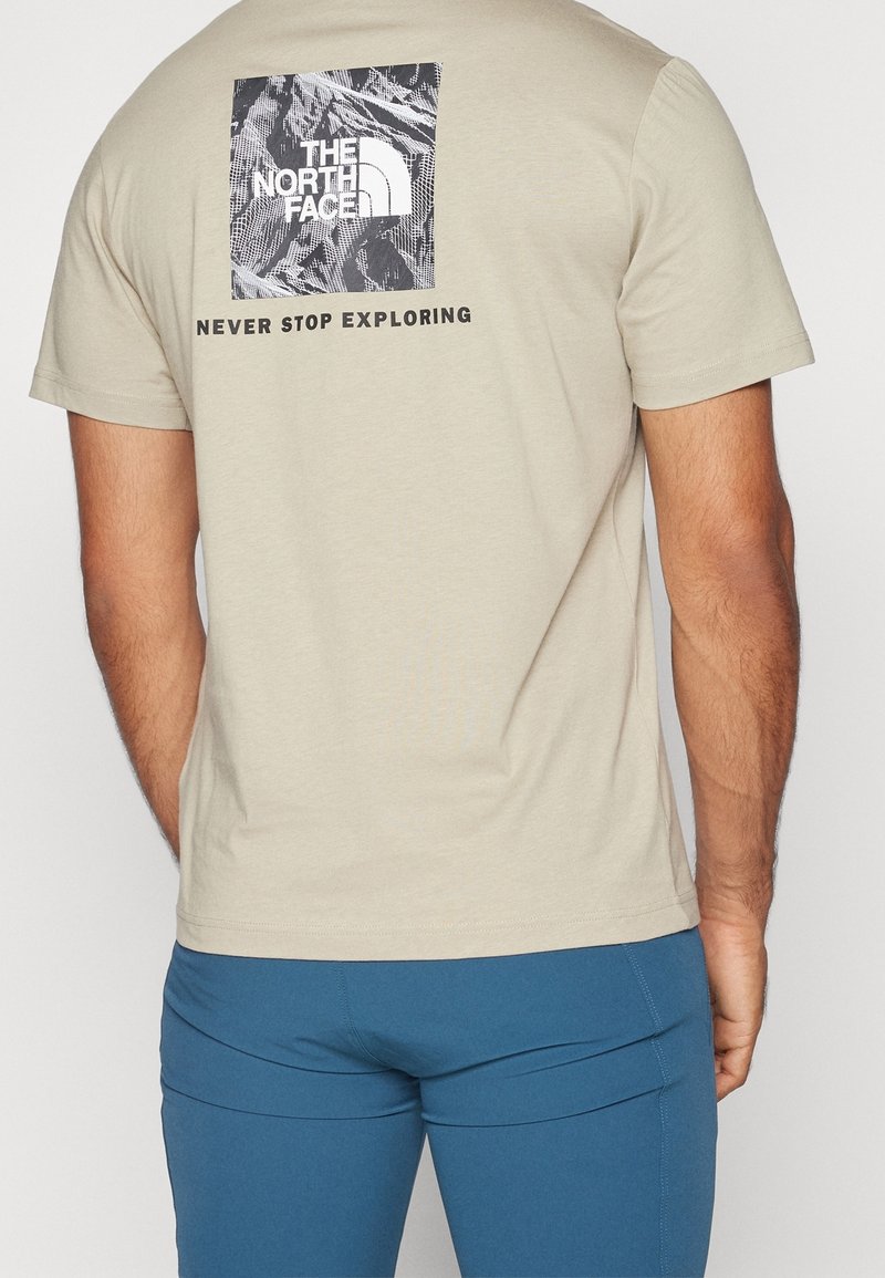 Muž v béžovém tričku s grafikou hory a logem "The North Face" s textem "Nikdy nepřestávejte prozkoumávat" na zadní straně, spárovaném s modrými kalhotami.