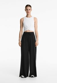 Weißes ärmelloses Crop-Top mit einem gedrehten Frontdetail, kombiniert mit schwarzen, weit geschnittenen Hosen mit gebundenem Bund und glatter Textur.