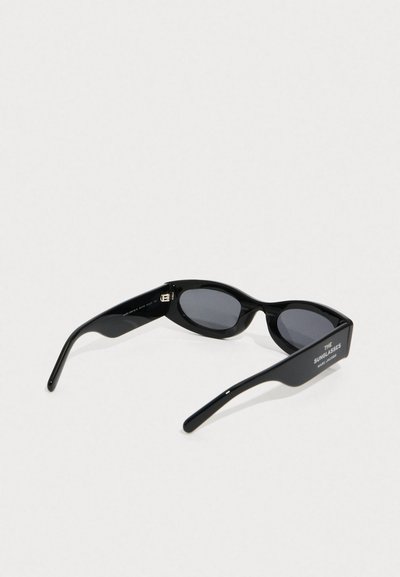 Occhiali da sole neri con montatura ovale, lenti scure, braccia larghe e "THE SUNGLASSES MARC JACOBS" stampato su un tempio.