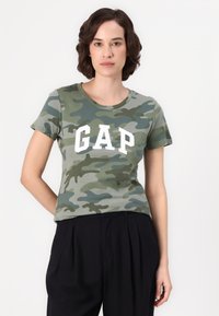 Grønn camo t-skjorte med hvit "GAP" tekst på forsiden; kort erm, rund hals, laget av myk bomull. Kombinert med svarte bukser.