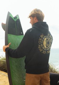 Sweat-shirt à capuche noir avec un graphisme doré, tenant une planche de surf verte avec une prise texturée et des détails noirs, sur fond côtier.