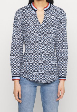 Kvinde iført en langærmet bluse med knapper, med blåt og hvidt geometrisk mønster samt rød, blå krave og manchetter, kombineret med lyseblå jeans.
