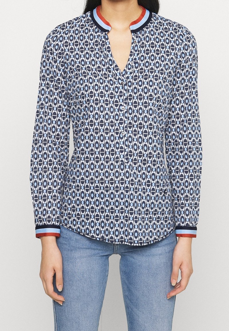 Femme portant un chemisier à manches longues boutonné, au motif géométrique bleu et blanc avec col et poignets rouges et bleus, associé à un jean bleu clair.