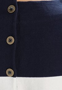 T-shirt tricoté avec des rayures horizontales bleu marine et blanches, présentant quatre boutons en ton laiton le long de la patte avant, et une texture lisse.