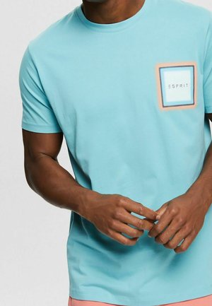 Uomo che indossa una maglietta azzurra a maniche corte con toppa quadrata con logo e la scritta "ESPRIT" sul petto sinistro, mani giunte davanti.