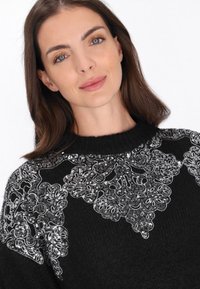 Zwarte gebreide trui met complexe zilveren sequins bloemenpatronen op de schouders, ronde hals en textuurdesign.