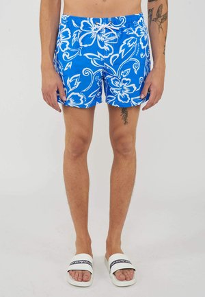 Uomo che indossa pantaloncini da bagno blu a fiori e sandali bianchi Emporio Armani, mostrando tatuaggi sull'avambraccio sinistro e sulla coscia destra.