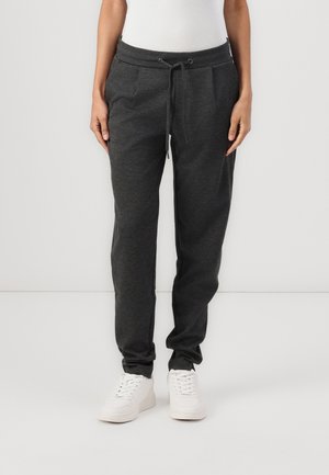 Persoon die donkere grijze joggingbroek met koord en witte sneakers draagt, staand tegen een effen lichte achtergrond.