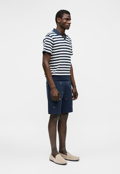 Hombre de pie vestido con un polo de manga corta a rayas azul marino y blanco, pantalones cortos azul marino con pequeñas estampas coloridas y zapatos beige sin cordones.