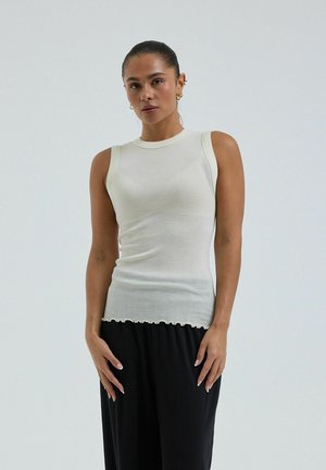 Vrouw draagt mouwloos roomkleurig geribd topje en losse zwarte broek, staat tegen een effen lichte achtergrond.