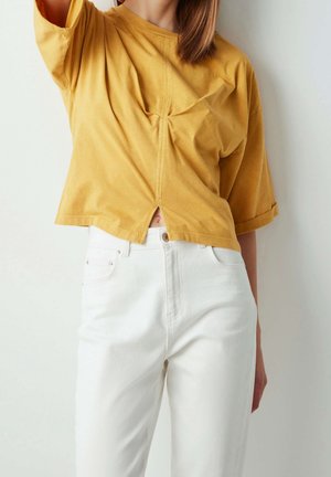 Femme portant un top court jaune moutarde avec une fente devant et un jean blanc taille haute, debout contre un mur uni.