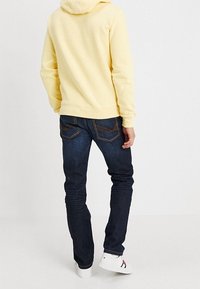 Sudadera amarilla con capucha, vaqueros azules oscuros con bolsillos traseros decorativos y zapatillas blancas con acentos en rojo y azul marino.