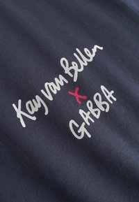 Námornícky modrý látka s bielym a červeným textom s nápisom "Kay van Bellen x GABBA." Mäkkejší povrch s uvoľneným dizajnom, zameriavajúcim sa na spoluprácu značiek.