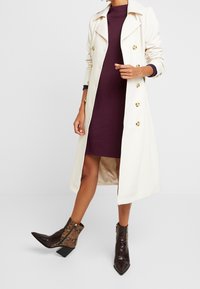 Cremefarbene Trenchcoat mit goldenen Knöpfen, über einem dunkelvioletten Kleid getragen. Ankle Boots mit Schlangenprint in Schwarz und Braun mit strukturierter Oberfläche.