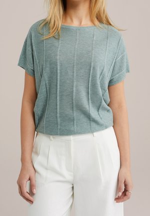 Femme portant un haut en maille vert clair à manches courtes avec des coutures verticales et un pantalon blanc plissé taille haute, debout devant un fond neutre.