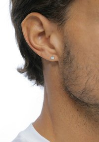 Quadratische Diamant-Ohrstecker aus silberfarbenem Metall, die an einem Ohrläppchen eines männlichen Ohrs mit kurzen, dunklen Haaren und leichtem Stoppelsitz befestigt sind.