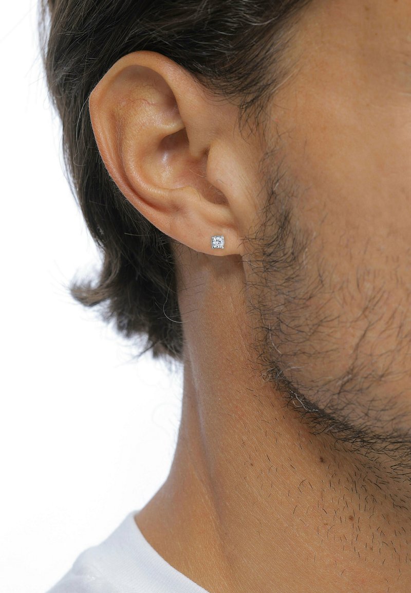 Quadratische Diamant-Ohrstecker aus silberfarbenem Metall, die an einem Ohrläppchen eines männlichen Ohrs mit kurzen, dunklen Haaren und leichtem Stoppelsitz befestigt sind.