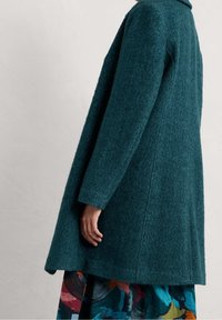 Cappotto di lana a spina di pesce color verde acqua indossato sopra una gonna a fantasia astratta colorata, su una persona in piedi contro un muro semplice di colore chiaro.