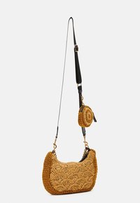 Tkaná slámová taška na rameno v zlatohnědé barvě s křivým tvarem, zdobená odnímatelnou kulatou kapsou. Černý popruh a zlaté kovové detaily.