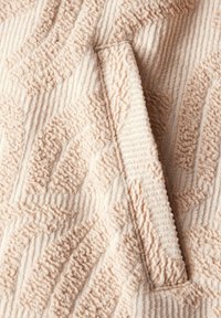 Tejido texturizado en color beige claro con un patrón en relieve. Incorpora un detalle de bolsillo discreto integrado en el diseño. Apariencia suave y táctil.