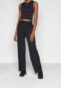 Osoba w czarnym bezrękawniku i luźnych czarnych spodniach, w czarnych sneakersach, stojąca na tle jasnego, gładkiego tła.