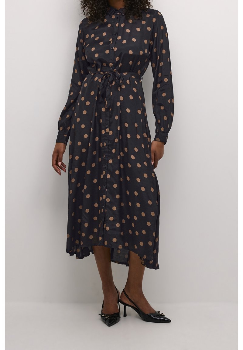 Femme portant une robe chemise midi noire à pois marron, manches longues, boutonnée devant, taille nouée et talons aiguilles noirs.