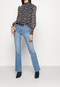 Black floral blouse with ruffled sleeves, light blue flared jeans, and black heeled boots. Accessorized with a small beige bag and black belt.