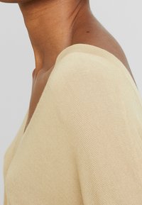 Haut en maille beige clair avec encolure en V et manches courtes. Présente une texture douce et une coupe ajustée, mettant en valeur des détails de couture soignés.