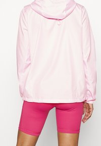 Jasnoróżowa kurtka z kapturem wykonana z gładkiego, lekkiego materiału, z elastycznymi mankietami i bocznymi kieszeniami; w komplecie z dopasowanymi różowymi szortami.