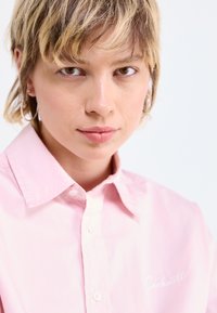 Camicia button-up rosa chiaro realizzata in tessuto liscio. Presenta un colletto classico, un logo ricamato sul lato sinistro del petto e una vestibilità strutturata.
