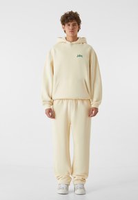 Sudadera con capucha color crema con bolsillo frontal y pequeño logo verde, combinada con pantalones de chándal de corte relajado a juego y zapatillas blancas.