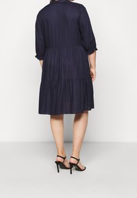 Vestido azul marino con un diseño de volantes, tela texturizada, mangas tres cuartos y puños ceñidos, combinado con zapatos de tacón negro con tiras.