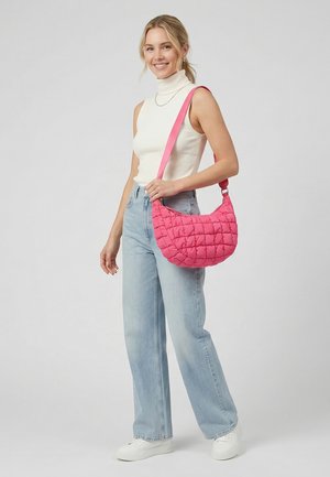 Mujer sonriendo, vestida con un jersey de cuello alto sin mangas blanco y jeans azul claro, sosteniendo un bolso acolchado rosa al hombro sobre un fondo liso.