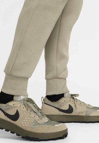 Beige Sportchuhe aus Mesh- und Wildledermaterialien, mit einem schwarzen Swoosh-Logo, strukturiertem Sohlen und Schnürdesign.